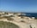 A coastal landscape featuring a rocky shoreline and expansive ocean view. White and beige buildings with red-tiled roofs are situated near the edge of the coast, surrounded by greenery and trees. The sky is clear and blue, enhancing the tranquil atmosphere of the scene.