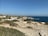 A coastal landscape featuring a rocky shoreline and expansive ocean view. White and beige buildings with red-tiled roofs are situated near the edge of the coast, surrounded by greenery and trees. The sky is clear and blue, enhancing the tranquil atmosphere of the scene.