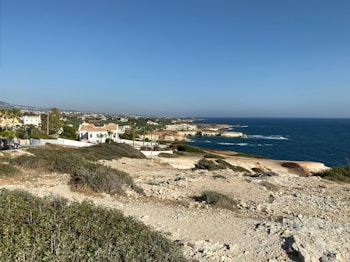A coastal landscape featuring a rocky shoreline and expansive ocean view. White and beige buildings with red-tiled roofs are situated near the edge of the coast, surrounded by greenery and trees. The sky is clear and blue, enhancing the tranquil atmosphere of the scene.