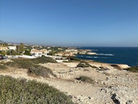 A coastal landscape featuring a rocky shoreline and expansive ocean view. White and beige buildings with red-tiled roofs are situated near the edge of the coast, surrounded by greenery and trees. The sky is clear and blue, enhancing the tranquil atmosphere of the scene.