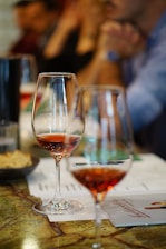 Two glasses of red wine are placed on a table with a marble surface. Several indistinct figures are in the background, potentially indicating social interaction. Papers and a menu are visible on the table, suggesting a dining or tasting setting.