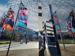 Colorful banners featuring various people hang on poles, set against a backdrop of a modern urban landscape. The scene includes reflective glass structures and geometric architecture under a partly cloudy sky.