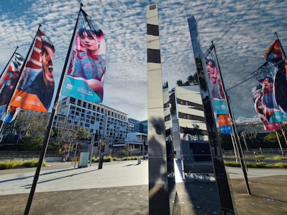 Colorful banners featuring various people hang on poles, set against a backdrop of a modern urban landscape. The scene includes reflective glass structures and geometric architecture under a partly cloudy sky.