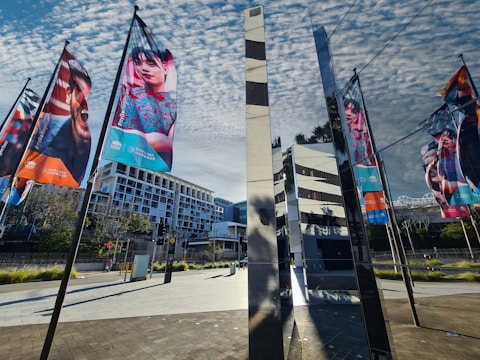 Colorful banners featuring various people hang on poles, set against a backdrop of a modern urban landscape. The scene includes reflective glass structures and geometric architecture under a partly cloudy sky.