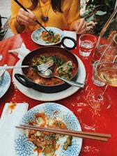 A dining scene featuring a red table with plates of food, primarily noodles with vegetables. A person with a yellow shirt is using chopsticks to eat from a plate. Various utensils, glasses of water, and a wine glass are present. The setting appears casual and relaxed, possibly outdoor, with some greenery in the background.
