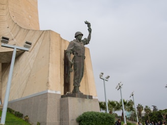 A large statue of a soldier stands prominently against a concrete structure. The soldier is wearing a helmet and uniform and is holding an object in one hand raised above his head. Surrounding the statue are modern pole lights, and there are palm trees and greenery at the base.