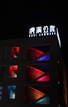 A building facade illuminated at night, showcasing a series of staircases behind square windows. Each level is lit with a different color, including red, pink, blue, and purple, creating a vibrant and artistic display. The sign on the building reads 'HUXI COMMUNE' in bright, white lettering.