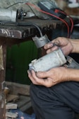 Hands are holding a worn electric motor part next to a workbench. There are visible wires and a multimeter in the background, suggesting electrical testing or repair work in progress.