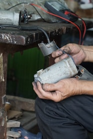 Hands are holding a worn electric motor part next to a workbench. There are visible wires and a multimeter in the background, suggesting electrical testing or repair work in progress.