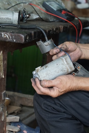 Hands are holding a worn electric motor part next to a workbench. There are visible wires and a multimeter in the background, suggesting electrical testing or repair work in progress.