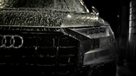 A close-up view of the front of a sleek, black car covered in water droplets, highlighting the car's luxury brand logo and stylish design. The scene is dimly lit, emphasizing the high-gloss finish and sleek headlight details.