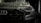 A close-up view of the front of a sleek, black car covered in water droplets, highlighting the car's luxury brand logo and stylish design. The scene is dimly lit, emphasizing the high-gloss finish and sleek headlight details.