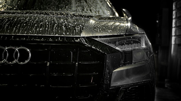 A close-up view of the front of a sleek, black car covered in water droplets, highlighting the car's luxury brand logo and stylish design. The scene is dimly lit, emphasizing the high-gloss finish and sleek headlight details.