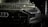 A close-up view of the front of a sleek, black car covered in water droplets, highlighting the car's luxury brand logo and stylish design. The scene is dimly lit, emphasizing the high-gloss finish and sleek headlight details.