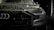A close-up view of the front of a sleek, black car covered in water droplets, highlighting the car's luxury brand logo and stylish design. The scene is dimly lit, emphasizing the high-gloss finish and sleek headlight details.