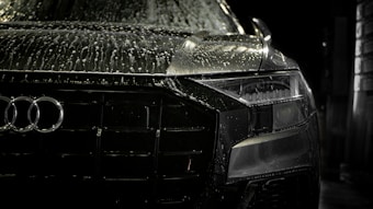 A close-up view of the front of a sleek, black car covered in water droplets, highlighting the car's luxury brand logo and stylish design. The scene is dimly lit, emphasizing the high-gloss finish and sleek headlight details.