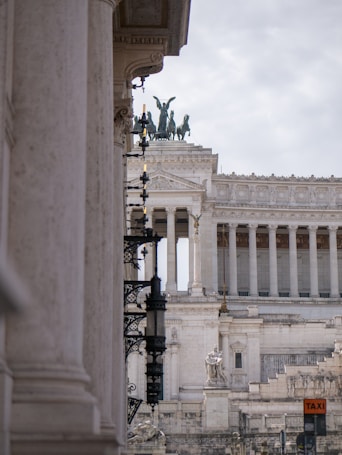 A majestic building with classical architecture, featuring tall columns and detailed sculptural elements. The top is adorned with a statue of a chariot and horses, symbolizing triumph. Ornate lamps line the side of the building, and a 'TAXI' sign is visible in the foreground. The sky is overcast, providing a neutral backdrop to the architectural details.