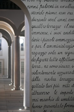 An ornate, archway with text inscribed on it stands in the foreground, featuring elegant, cursive handwriting. Behind it, a series of architectural arches with columns are softly illuminated. The light creates a serene atmosphere, highlighting the classic design elements.