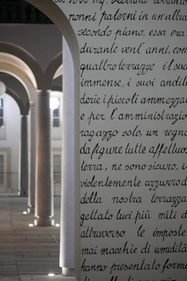 An ornate, archway with text inscribed on it stands in the foreground, featuring elegant, cursive handwriting. Behind it, a series of architectural arches with columns are softly illuminated. The light creates a serene atmosphere, highlighting the classic design elements.