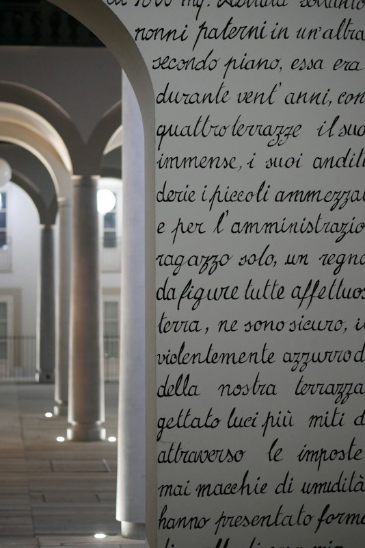 An ornate, archway with text inscribed on it stands in the foreground, featuring elegant, cursive handwriting. Behind it, a series of architectural arches with columns are softly illuminated. The light creates a serene atmosphere, highlighting the classic design elements.