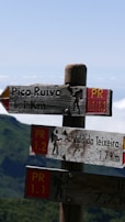Wooden hiking trail signs indicate directions to Pico Ruivo and Achada do Teixeira with distances in kilometers. The signs have a rustic appearance, featuring hiking icons and PR trail numbers. The background displays a mountainous landscape with lush greenery under a clear blue sky.