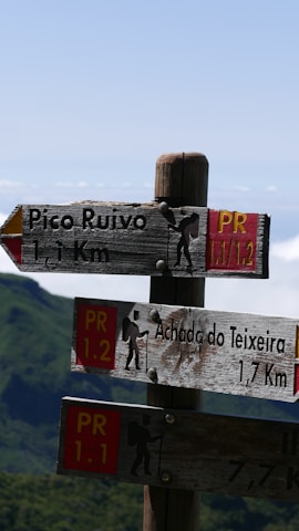 Wooden hiking trail signs indicate directions to Pico Ruivo and Achada do Teixeira with distances in kilometers. The signs have a rustic appearance, featuring hiking icons and PR trail numbers. The background displays a mountainous landscape with lush greenery under a clear blue sky.