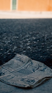 A vintage newspaper lies crumpled on a dark, textured asphalt surface. The background features a softly focused orange wall with a green window, adding contrast to the foreground.