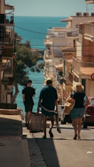 A bustling street descends towards a scenic view of the sea, framed by buildings on either side. The warm sunlight casts shadows, enhancing the coastal ambiance. People are walking down the street, some carrying luggage, suggesting a travel or vacation setting.