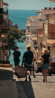 A bustling street descends towards a scenic view of the sea, framed by buildings on either side. The warm sunlight casts shadows, enhancing the coastal ambiance. People are walking down the street, some carrying luggage, suggesting a travel or vacation setting.