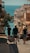 A bustling street descends towards a scenic view of the sea, framed by buildings on either side. The warm sunlight casts shadows, enhancing the coastal ambiance. People are walking down the street, some carrying luggage, suggesting a travel or vacation setting.