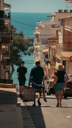 A bustling street descends towards a scenic view of the sea, framed by buildings on either side. The warm sunlight casts shadows, enhancing the coastal ambiance. People are walking down the street, some carrying luggage, suggesting a travel or vacation setting.