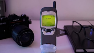 A vintage mobile phone with a small antenna is displayed prominently. Next to it, there is an older model film camera with a large lens. Resting on a table is a pair of reading glasses and a closed notebook, suggesting a setting that blends technology and personal organization.