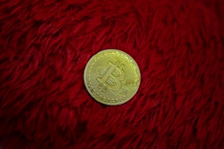A gold Bitcoin representing a cryptocurrency is placed on a textured red surface, possibly a furry or shaggy fabric.