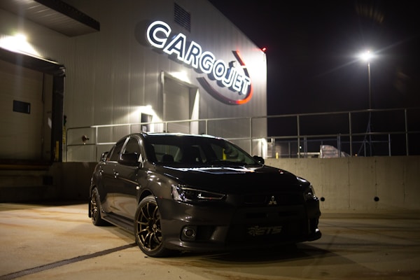 A sleek black car is parked at night in front of a building with bright signage displaying the word 'Cargojet'. The scene is illuminated by artificial lights creating reflections on the car's surface and casting shadows on the ground.