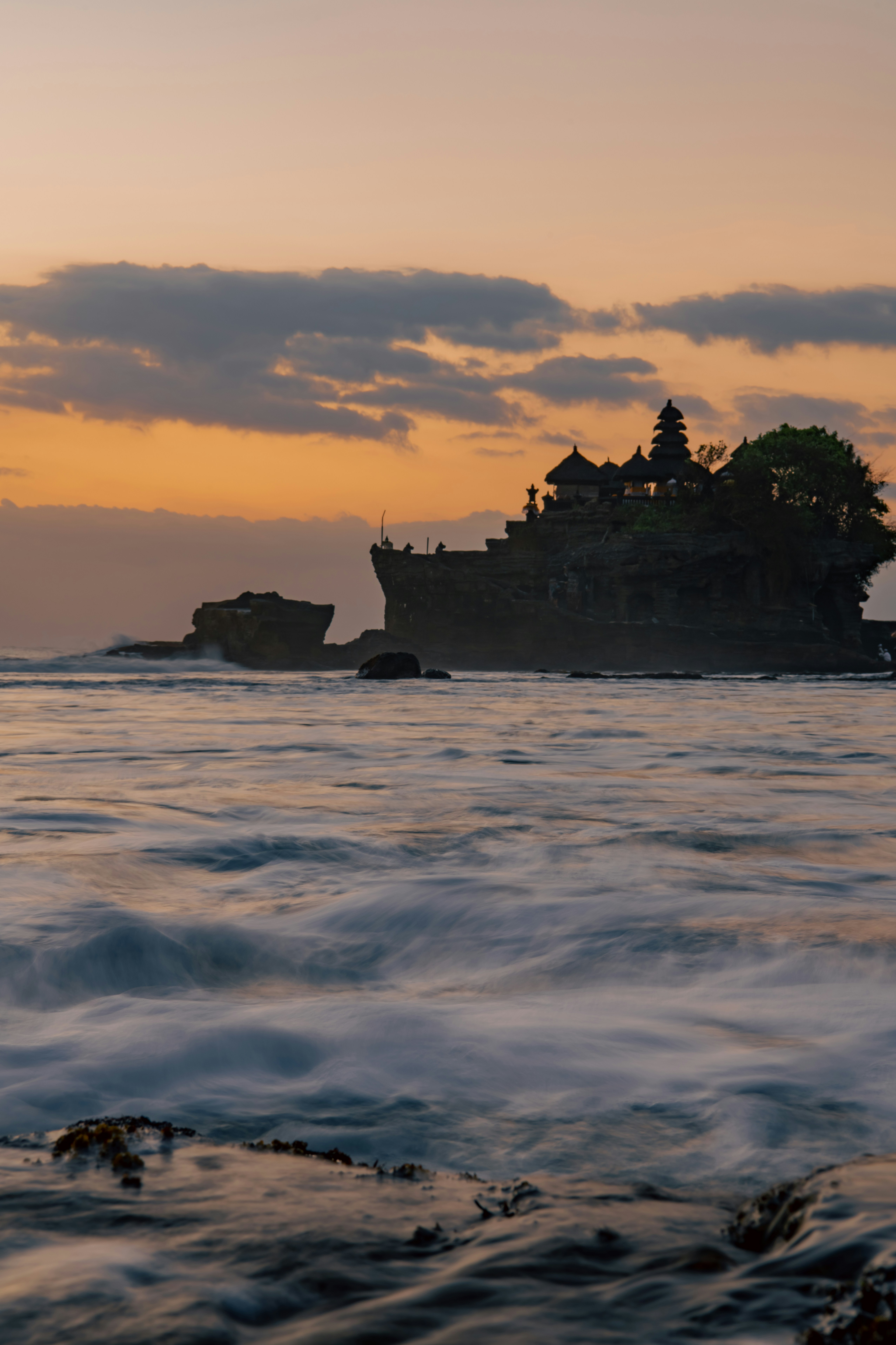 Templo Tanah Lot en Bali sobre roca volcánica al atardecer, olas del océano índico rompiendo, cielo naranja