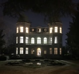 A large, elegant mansion illuminated from within against a nighttime backdrop. Two cylindrical towers flank the main structure, which features multiple arched windows and a central doorway. The foreground includes a garden path and reflection pool, with surrounding trees framing the scene.