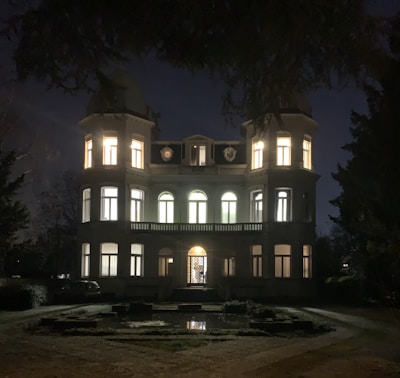 A large, elegant mansion illuminated from within against a nighttime backdrop. Two cylindrical towers flank the main structure, which features multiple arched windows and a central doorway. The foreground includes a garden path and reflection pool, with surrounding trees framing the scene.
