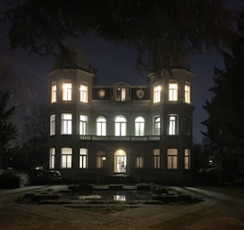 A large, elegant mansion illuminated from within against a nighttime backdrop. Two cylindrical towers flank the main structure, which features multiple arched windows and a central doorway. The foreground includes a garden path and reflection pool, with surrounding trees framing the scene.