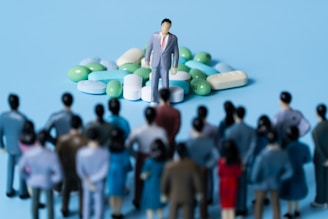 A crowd of miniature figurines dressed in business attire stands facing a single figure placed on a pile of assorted pills and capsules. The scene appears to be set against a light blue background.