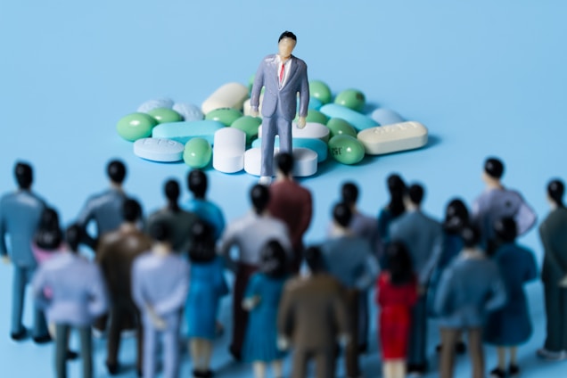A crowd of miniature figurines dressed in business attire stands facing a single figure placed on a pile of assorted pills and capsules. The scene appears to be set against a light blue background.
