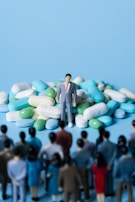 A miniature figure of a man in a suit stands on a large pile of assorted pills. In the foreground, several smaller figures appear to be looking towards the man and the pile of pills. The background is a solid light blue color.