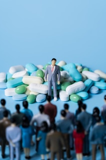 A miniature figure of a man in a suit stands on a large pile of assorted pills. In the foreground, several smaller figures appear to be looking towards the man and the pile of pills. The background is a solid light blue color.