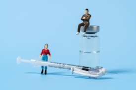 Two miniature figures positioned with medical items, one figure leans against a syringe while another sits atop a glass vial containing liquid. The figures appear to be representing people in a conceptual display against a light blue background.