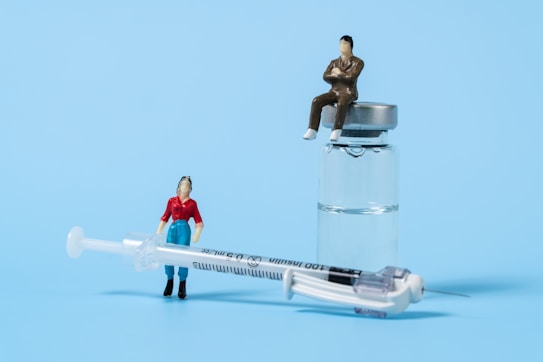Two miniature figures positioned with medical items, one figure leans against a syringe while another sits atop a glass vial containing liquid. The figures appear to be representing people in a conceptual display against a light blue background.