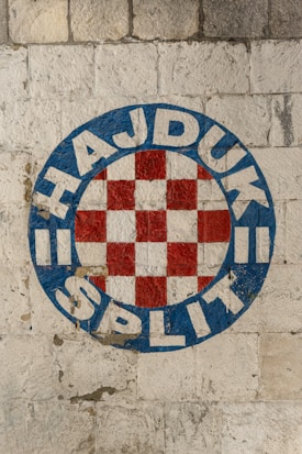 A logo featuring a circular emblem with a red and white checkered pattern in the center, surrounded by a blue border. The words 'HAJDUK SPLIT' are written in bold white letters within the blue border. The emblem appears painted on a textured stone wall.