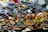 A variety of colorful sandals are displayed on a table. The sandals feature intricate designs and patterns, with straps in vibrant colors such as orange, blue, green, and yellow. In the background, there are darker-toned leather shoes in less vivid hues.
