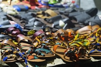A variety of colorful sandals are displayed on a table. The sandals feature intricate designs and patterns, with straps in vibrant colors such as orange, blue, green, and yellow. In the background, there are darker-toned leather shoes in less vivid hues.