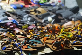 A variety of colorful sandals are displayed on a table. The sandals feature intricate designs and patterns, with straps in vibrant colors such as orange, blue, green, and yellow. In the background, there are darker-toned leather shoes in less vivid hues.