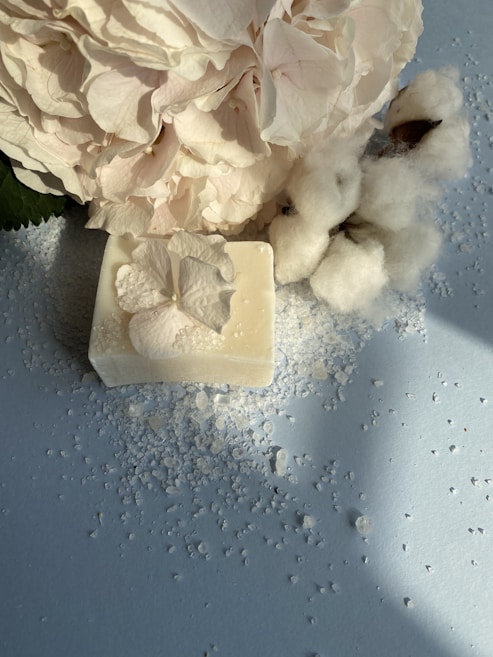 A bar of soap is adorned with a delicate white flower on its surface, accompanied by scattered salt crystals. Nearby, there is a cotton plant with fluffy white bolls, contrasting against the soft blue background. A large, pale hydrangea flower is positioned artistically, adding an elegant touch.