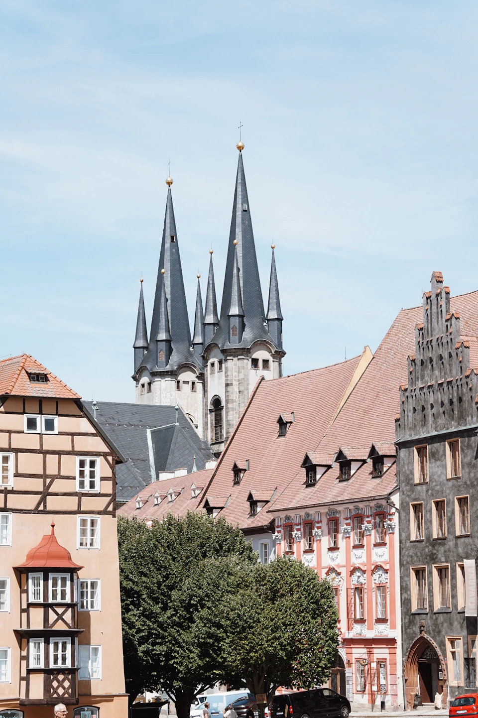 The image depicts a picturesque European town with a row of buildings featuring traditional architecture, including a church with two tall spires and a clock tower. The buildings are painted in various colors, creating a vibrant and diverse scene. In front of the church, there is a tree that adds a touch of nature to the urban landscape.

The sky above is clear blue, providing a beautiful backdrop for the colorful buildings below. There are several cars parked on the street, indicating that this town is accessible by road transportation. The image does not contain any discernible text or countable objects, and there are no people visible in the scene.